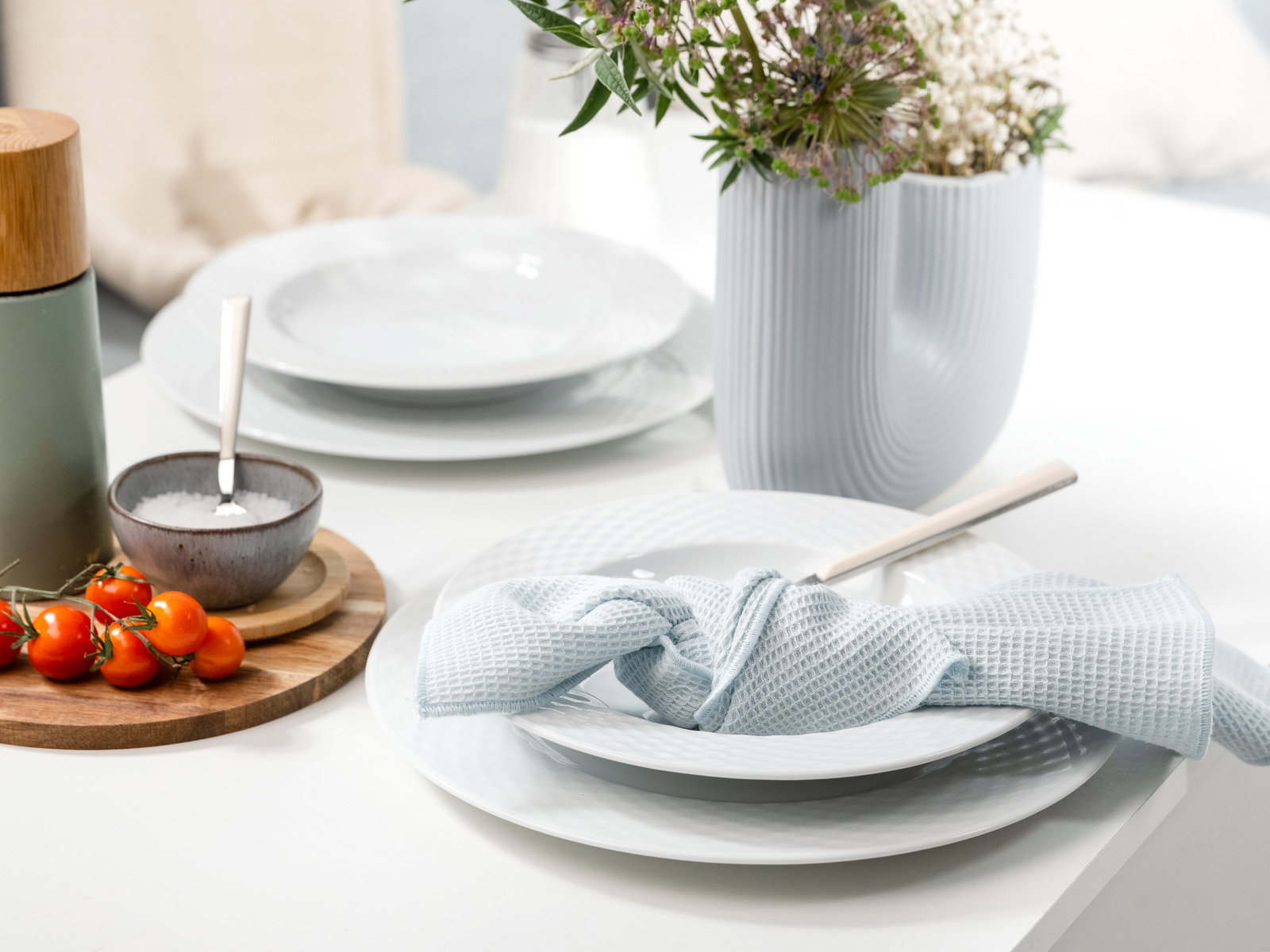 Elegantes Tafelservice mit weißen Tellern, blauer Serviette und Besteck, dekoriert mit Blumen und Kirschtomaten, seitliche Perspektive.