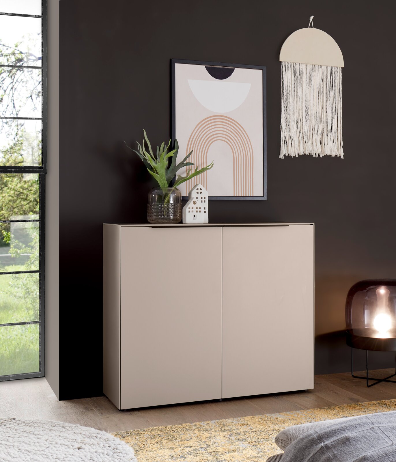 Voleo Kommode ROSSO Beiges Sideboard mit zwei Türen in moderner, minimalistischer Optik, frontal aus leicht schräger Perspektive fotografiert, dekoriert mit Glasvase und gerahmtem Poster vor dunkler Wand.