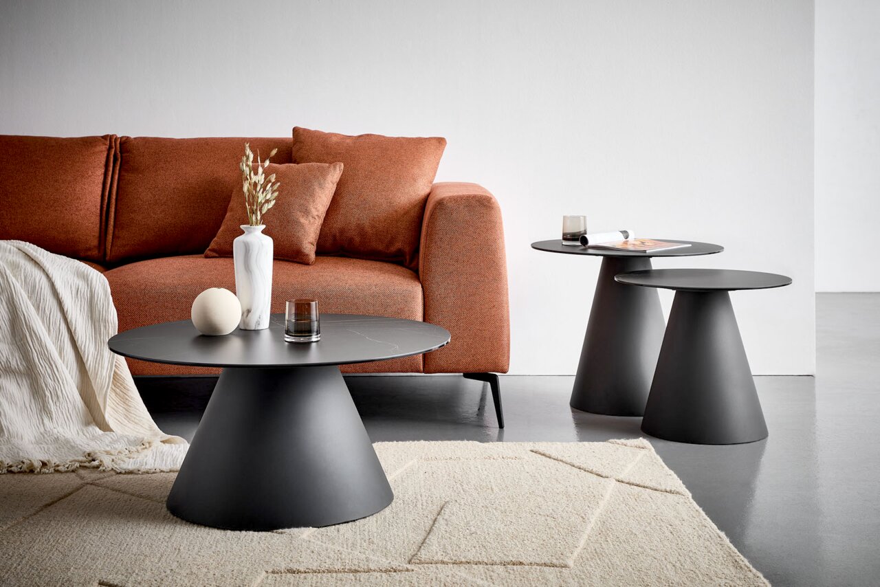 Novel Beistelltisch COMO Frontale Wohnraumansicht eines modernen dunkelgrauen Couchtisch-Sets mit runden Tischplatten und konischen Sockeln vor einem rostfarbenen Sofa.