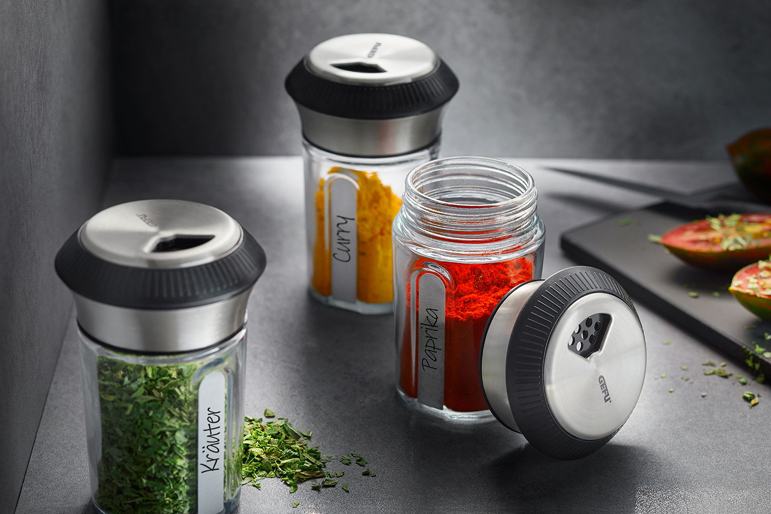 Drei Gewürz- und Kräuterstreuer mit Edelstahlkappen, gefüllt mit Curry, Paprika und Kräutern, auf einer Küchenarbeitsplatte. Perspektive von schräg oben.