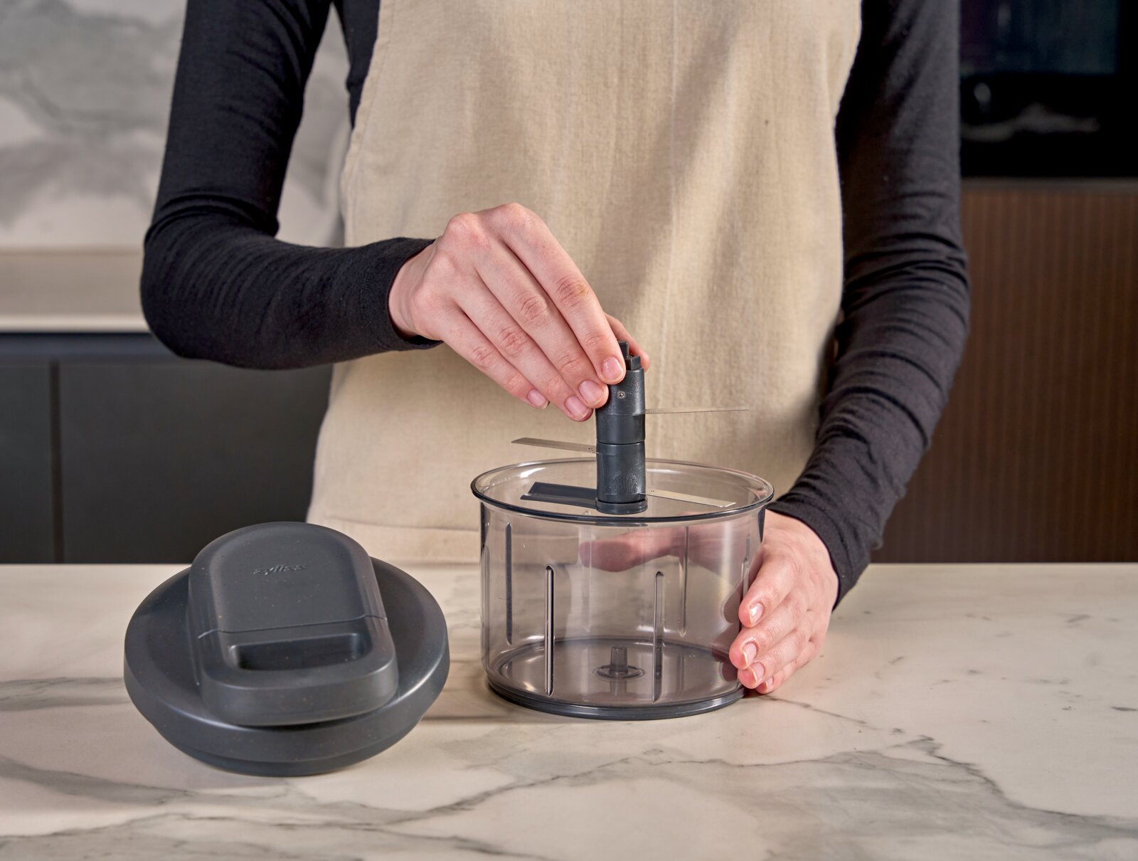 Keukenaccessoire: transparante hakmolen/foodprocessor-kom met mesunit en los deksel op een marmeren aanrecht, vastgehouden door een persoon in schort, frontaal aanzicht op werkhoogte