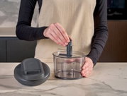 Keukenaccessoire: transparante hakmolen/foodprocessor-kom met mesunit en los deksel op een marmeren aanrecht, vastgehouden door een persoon in schort, frontaal aanzicht op werkhoogte