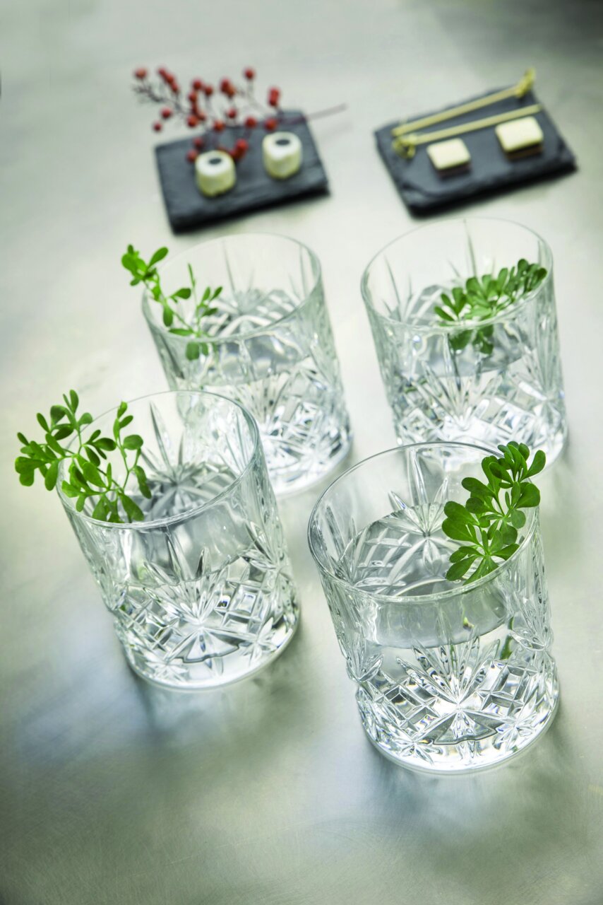 Vier Whiskygläser aus Kristallglas mit dekorativem Schliff, von oben fotografiert, mit grünen Zweigen als Dekoration.