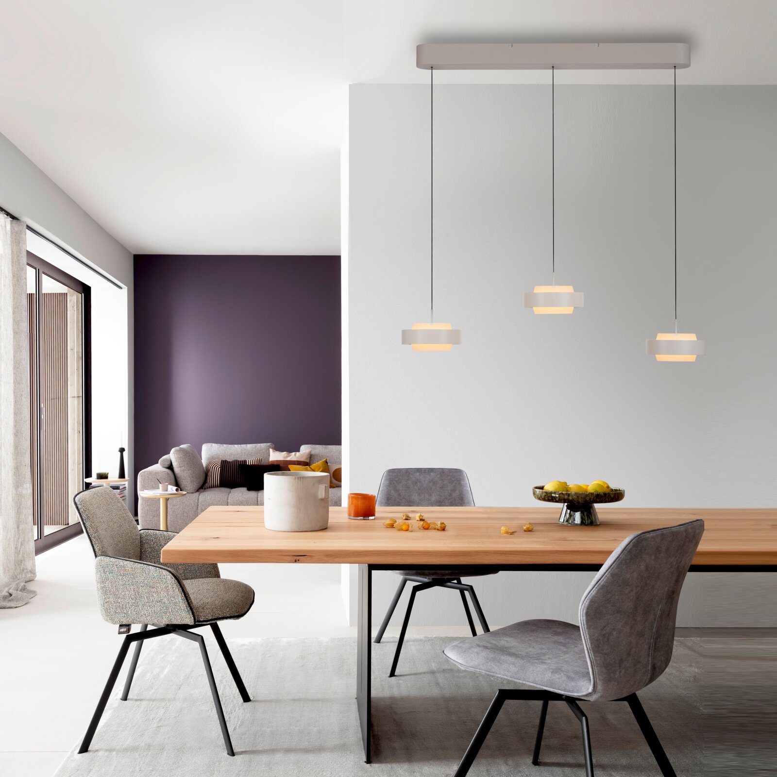 Moderne hanglamp met drie lampen, voorzien van drie kleine, ronde lampenkappen in beige, aan een zwarte kabel en met een langwerpige plafondbevestiging boven een eettafel; vooraanzicht van de ruimte