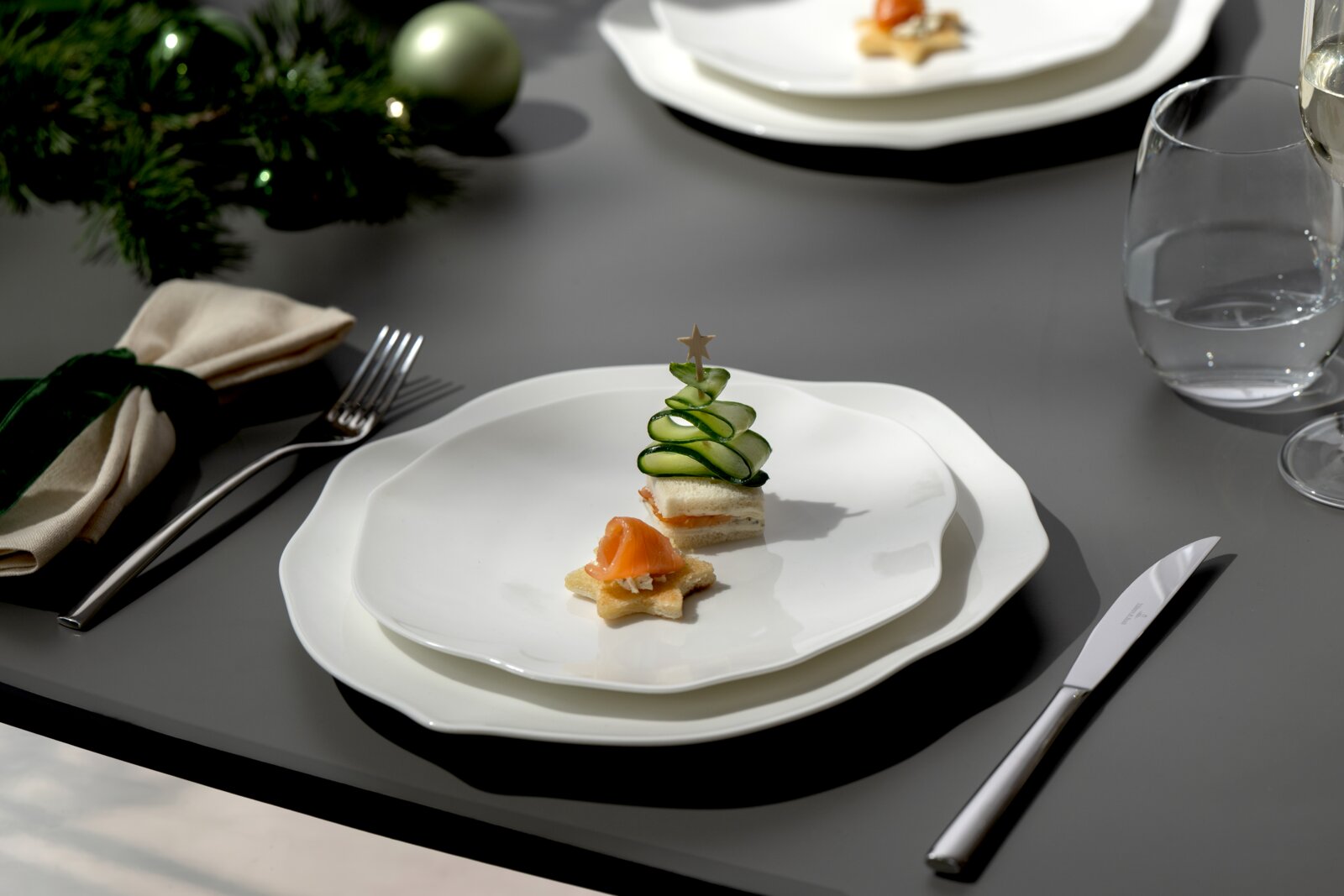 Weiße, wellenförmige Teller auf grauem Tisch mit weihnachtlichen Häppchen (Gurkenbaum und Lachs auf Stern-Cracker), gedecktem Besteck und Glas, aus leicht schräger Draufsicht