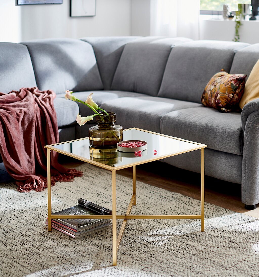 Quadratischer Couchtisch mit Spiegelglasplatte und goldfarbenem Metallgestell im Wohnzimmer vor grauem Ecksofa, Perspektive: leicht schräg von vorne auf Tischhöhe