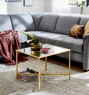 Quadratischer Couchtisch mit Spiegelglasplatte und goldfarbenem Metallgestell im Wohnzimmer vor grauem Ecksofa, Perspektive: leicht schräg von vorne auf Tischhöhe