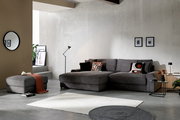 Modernes Wohnzimmer mit grauem Cord-Ecksofa mit Ottomane, passenden Kissen und Hocker, aufgenommen in frontaler, leicht seitlicher Perspektive.