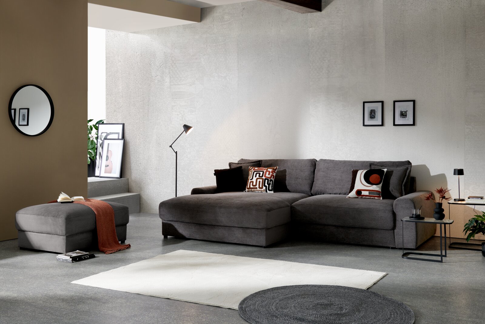 Modernes Wohnzimmer mit grauem Cord-Ecksofa mit Ottomane, passenden Kissen und Hocker, aufgenommen in frontaler, leicht seitlicher Perspektive.