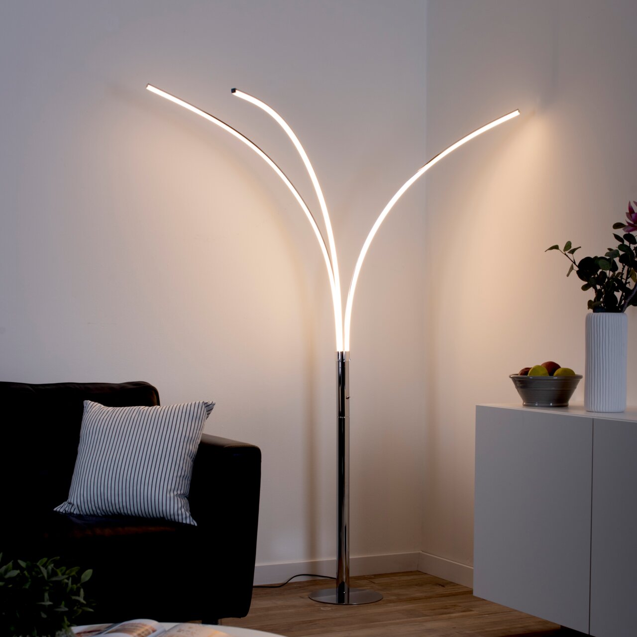 Moderne Stehleuchte aus Chrom mit geschwungenen LED-Lichtarmen, in einer gemütlichen Wohnzimmerumgebung, seitliche Perspektive