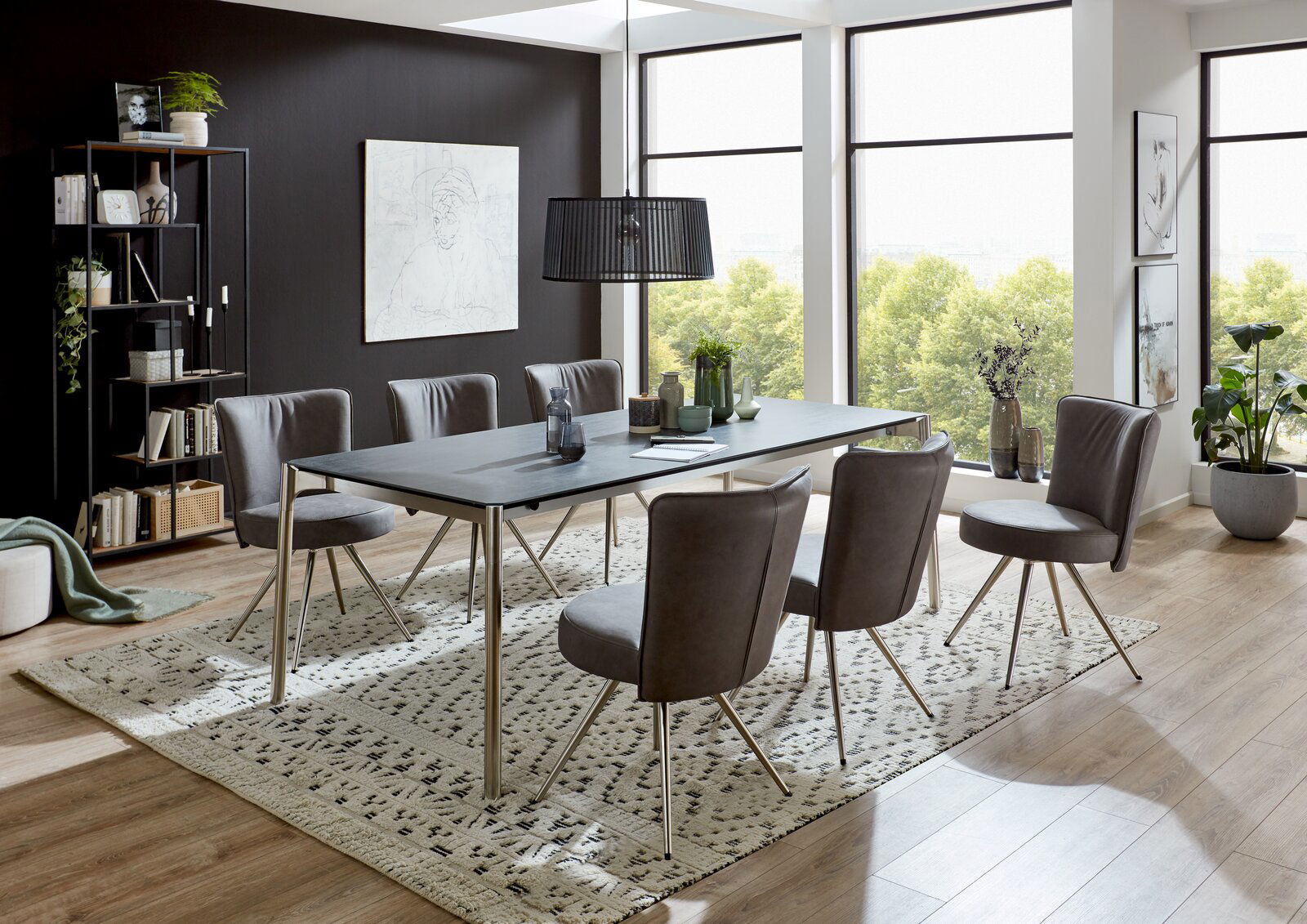Moderner Esstisch mit Metallbeinen und dunkler Tischplatte, umgeben von sechs grauen Stühlen in einem hellen Raum mit großen Fenstern, seitliche Perspektive.