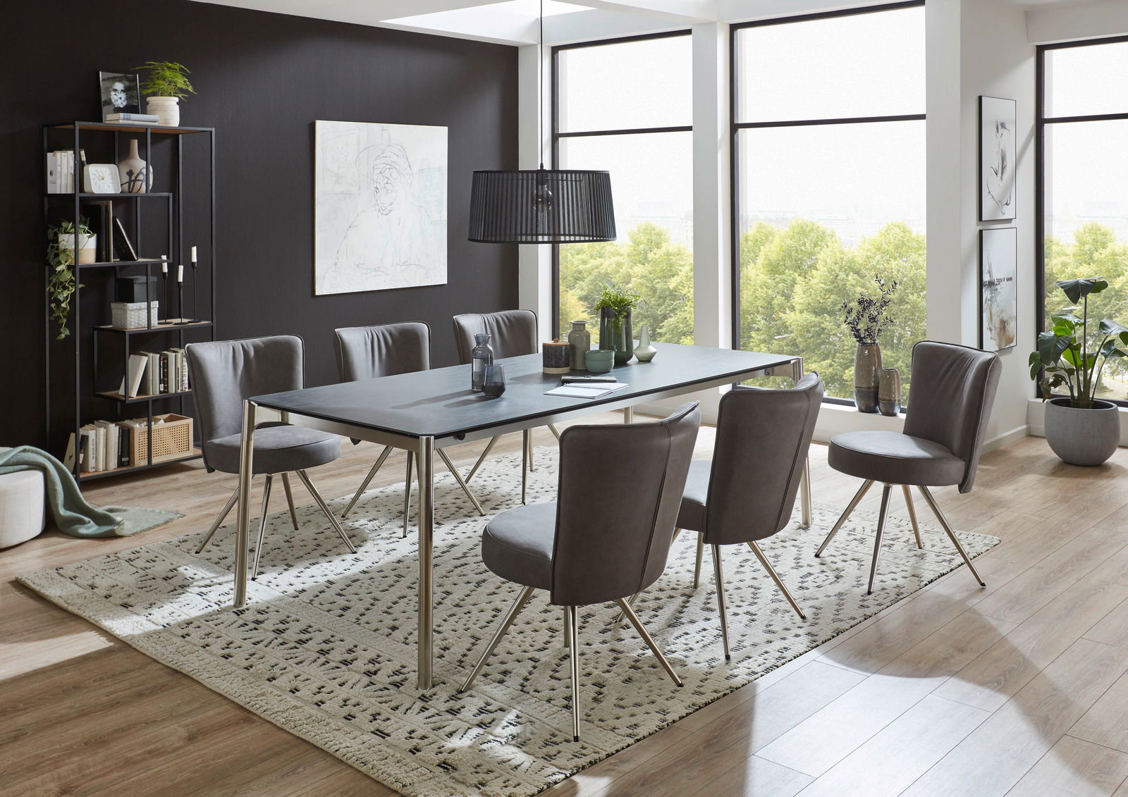 Moderner Esstisch mit Metallbeinen und dunkler Tischplatte, umgeben von sechs grauen Stühlen in einem hellen Raum mit großen Fenstern, seitliche Perspektive.