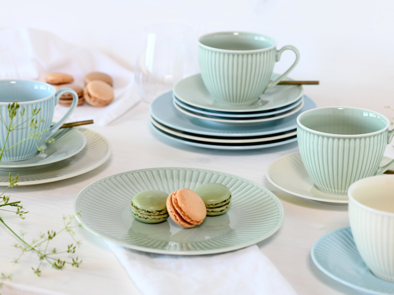 Pastellfarbenes geripptes Geschirr-Set mit Tassen, Untertassen und Tellern in Mint und Hellblau, arrangiert auf weißem Tisch mit Macarons; Perspektive: leicht erhöhte Schrägansicht von vorn.