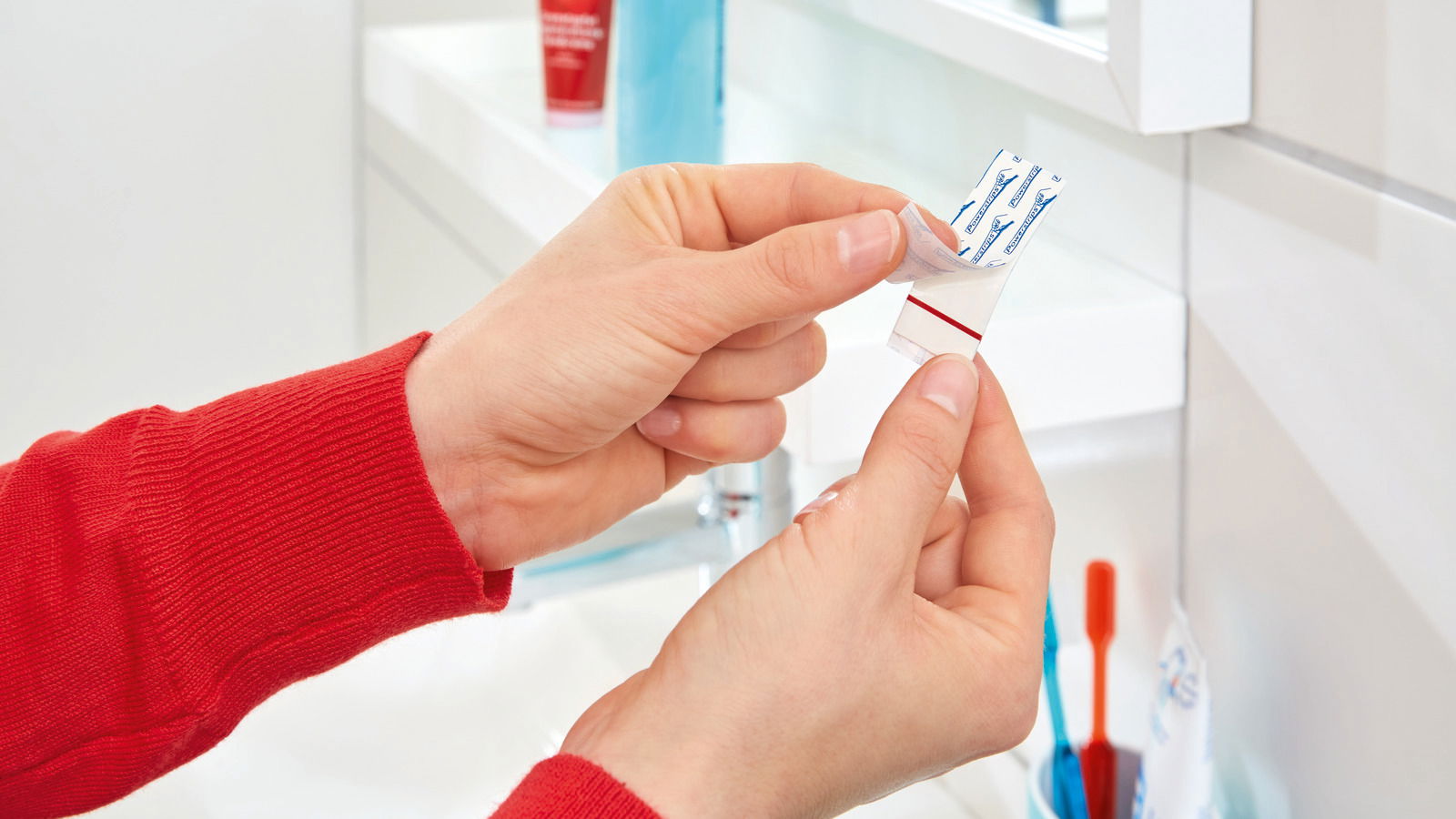 Nahaufnahme von Händen, die einen tesa Powerstrip in einem Badezimmer halten, mit einem Spiegel und Zahnbürsten im Hintergrund.