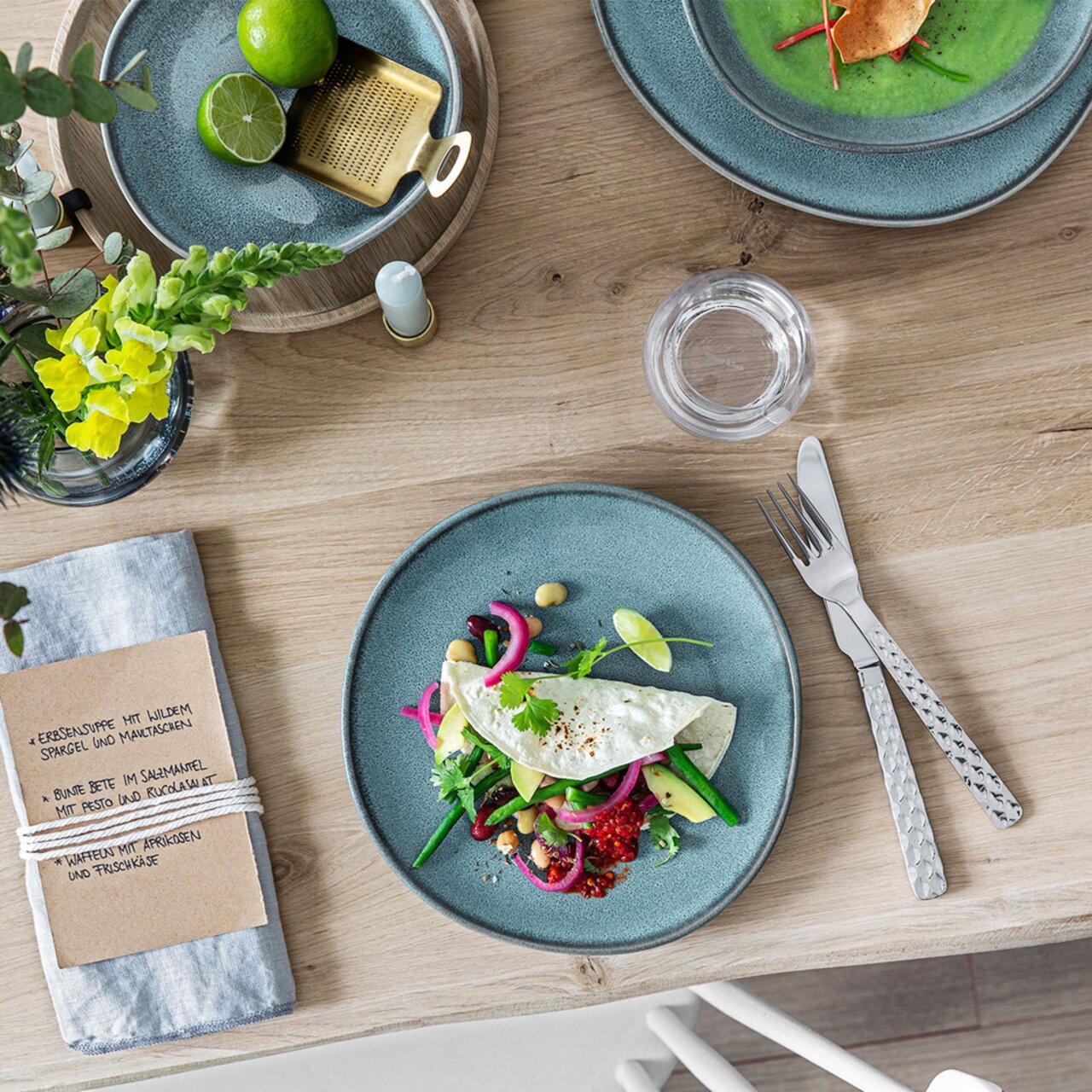 Like by Villeroy & Boch Schale flach LAVE GLACE Vogelperspektive auf einen gedeckten Tisch mit einer türkisfarbenen Obstschale, gefüllt mit Limetten, und einem Teller mit einer bunten Speise.