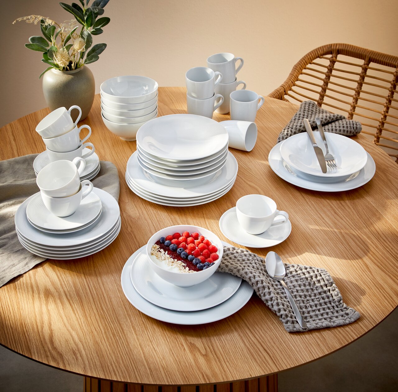 Kombiservice Beat 42-tlg. auf einem Holztisch, bestehend aus weißen Tellern, Tassen und Schalen, mit einer modernen Tischdekoration aus einer Vase und Besteck. Perspektive von oben.