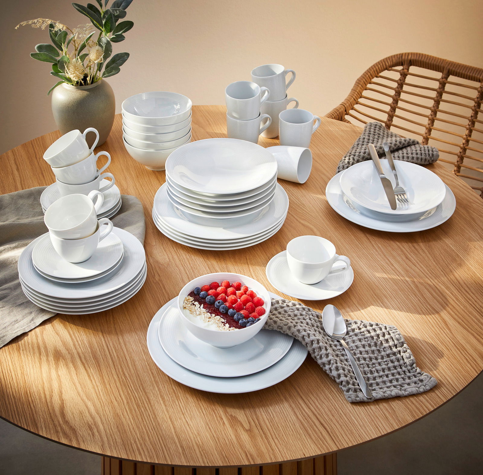 Kombiservice Beat 42-tlg. auf einem Holztisch, bestehend aus weißen Tellern, Tassen und Schalen, mit einer modernen Tischdekoration aus einer Vase und Besteck. Perspektive von oben.