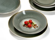 Grijs-groen keramisch servies met diepe kom op een bord en gestapelde borden op witte achtergrond, met kleine tomaatjes en bonen als styling; gefotografeerd in schuin bovenaanzicht.