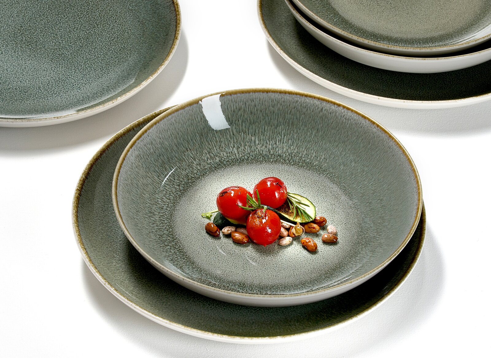 Grijs-groen keramisch servies met diepe kom op een bord en gestapelde borden op witte achtergrond, met kleine tomaatjes en bonen als styling; gefotografeerd in schuin bovenaanzicht.