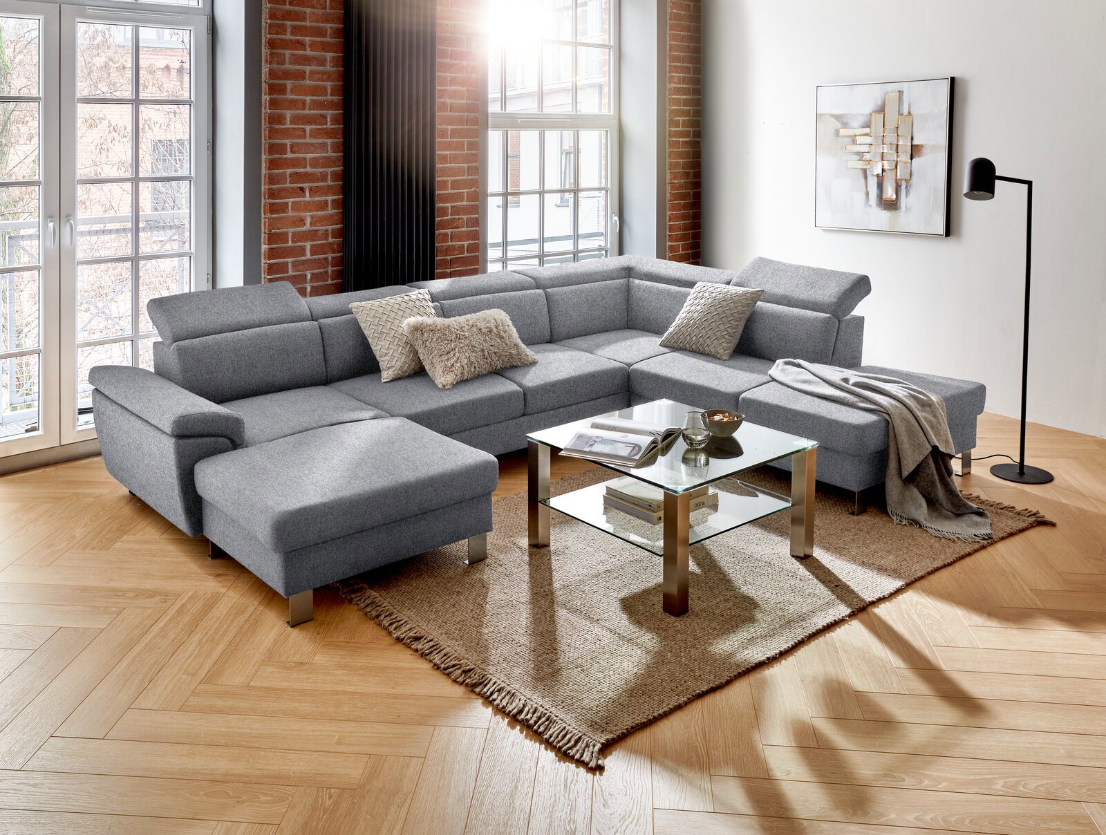 Graues Ecksofa in U-Form in einem modernen Wohnzimmer mit Holzboden, Glas-Couchtisch und großen Fenstern, seitliche Perspektive.