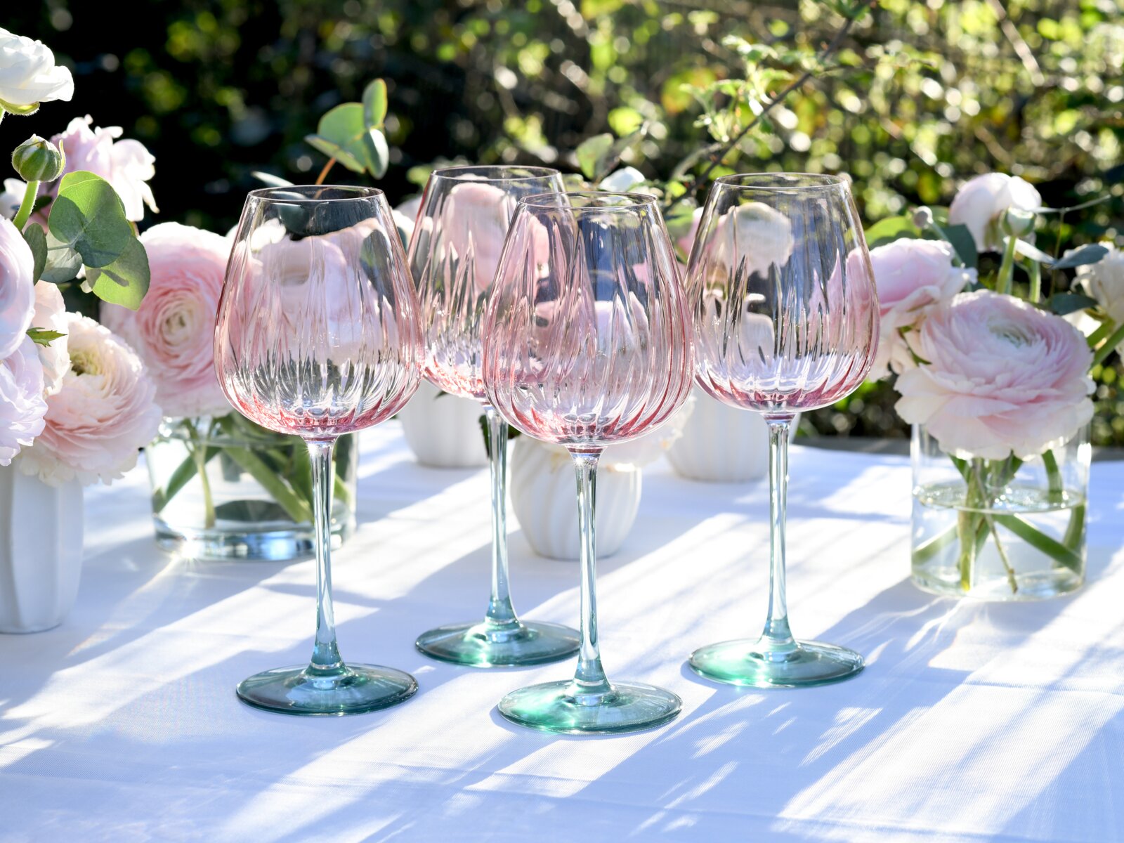 Vier elegante Weingläser aus klarem Glas mit zart roséfarbenem Verlauf und geriffelter Struktur, auf weißer Tischdecke zwischen rosa Blumenvasen im Sonnenlicht, frontal aus leicht niedriger Perspektive aufgenommen