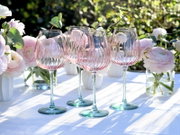 Vier elegante Weingläser aus klarem Glas mit zart roséfarbenem Verlauf und geriffelter Struktur, auf weißer Tischdecke zwischen rosa Blumenvasen im Sonnenlicht, frontal aus leicht niedriger Perspektive aufgenommen