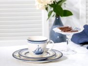 Weiß-blaues Porzellan-Kaffeegedeck mit Tasse und Untertasse im maritimen Segelschiff-Design auf Tischdecke, seitliche Perspektive auf Augenhöhe; im Hintergrund Glas-Etagere mit Schokoladenstücken und blaue Vase mit Blumen