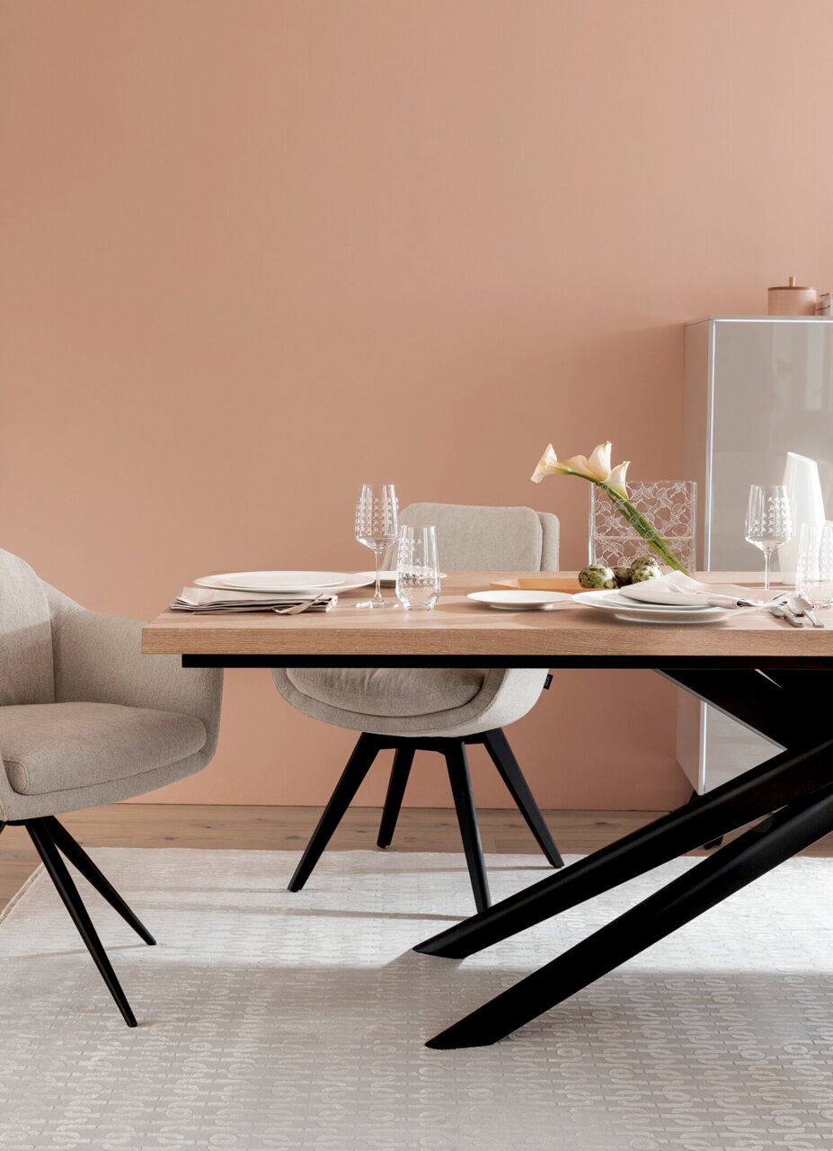 Moderner Esstisch mit Holzplatte und schwarzen Metallbeinen, umgeben von beigen Stühlen, gedeckt mit elegantem Geschirr und Gläsern, seitliche Perspektive.