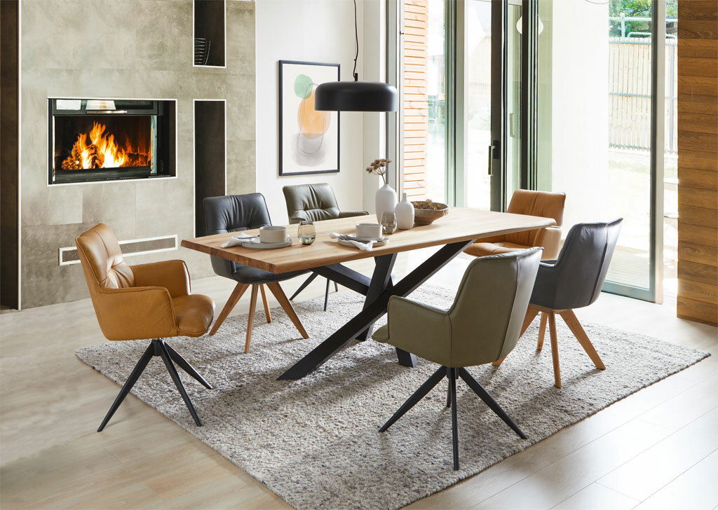 Moderner Esstisch mit Holzplatte und schwarzen Metallbeinen, umgeben von verschiedenen gepolsterten Stühlen in einem stilvollen Esszimmer mit Kamin. Perspektive: seitliche Ansicht.