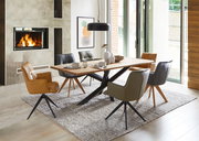 Moderner Esstisch mit Holzplatte und schwarzen Metallbeinen, umgeben von verschiedenen gepolsterten Stühlen in einem stilvollen Esszimmer mit Kamin. Perspektive: seitliche Ansicht.