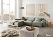Moderne grüne Ecksofa-Landschaft in einem hellen Wohnzimmer mit großen Fenstern, einem flauschigen Teppich und einem Beistelltisch. Perspektive von vorne links.