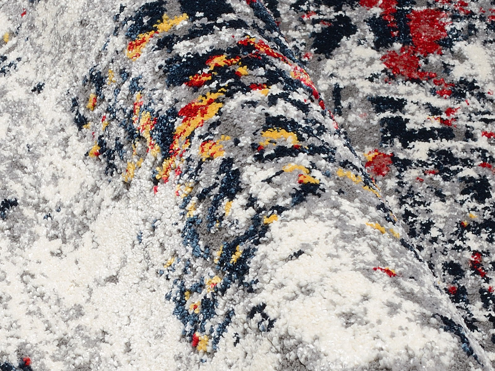 Nahaufnahme eines Designer-Teppichs mit abstraktem Muster in Grau, Weiß, Rot, Gelb und Blau. Die Perspektive zeigt eine seitliche Ansicht der Textur und Farben.