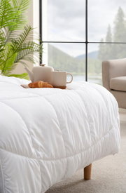 Gemütliche weiße Decke auf einem Bett mit Teekanne, Tasse und Croissant, im Hintergrund ein großes Fenster mit Blick auf die Natur, seitliche Perspektive.