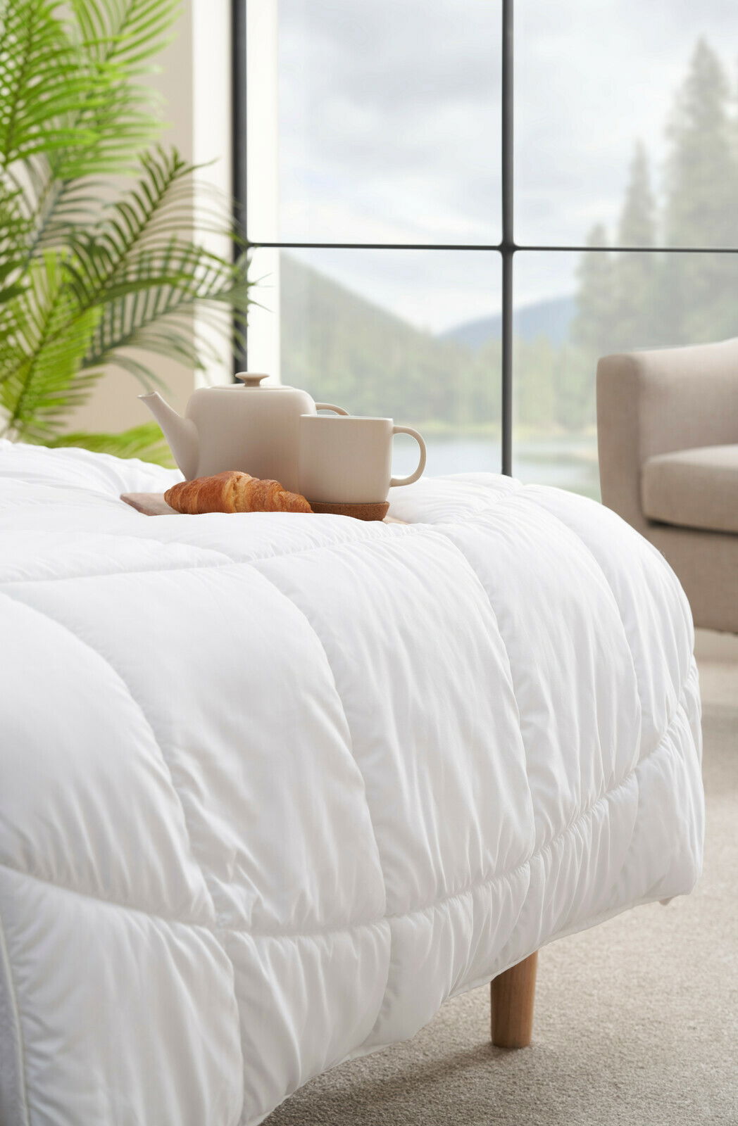 Gemütliche weiße Decke auf einem Bett mit Teekanne, Tasse und Croissant, im Hintergrund ein großes Fenster mit Blick auf die Natur, seitliche Perspektive.