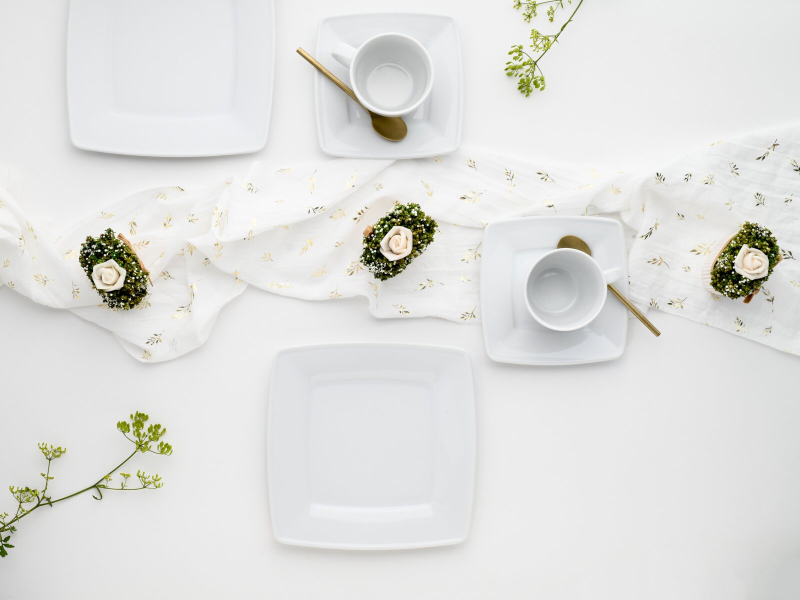 Elegantes weißes, quadratisches Geschirr-Set mit Tellern und Tassen auf hellem Tisch, goldene Löffel und florale Deko; Draufsicht von oben (Flatlay).