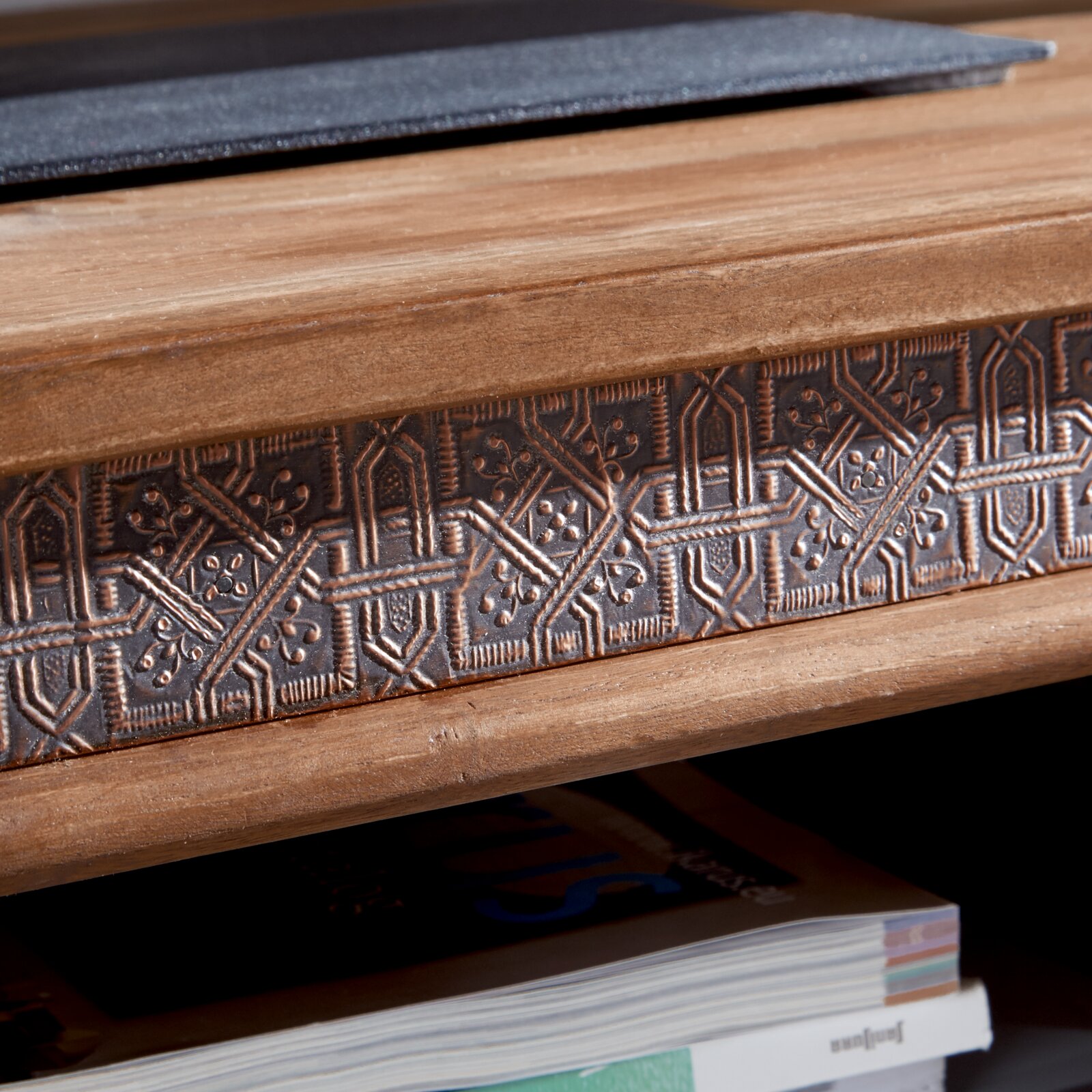 Close-up van een tv-meubel met een decoratief, bewerkt houten detail en een tijdschrift op de plank eronder, gezien vanuit de zijkant.