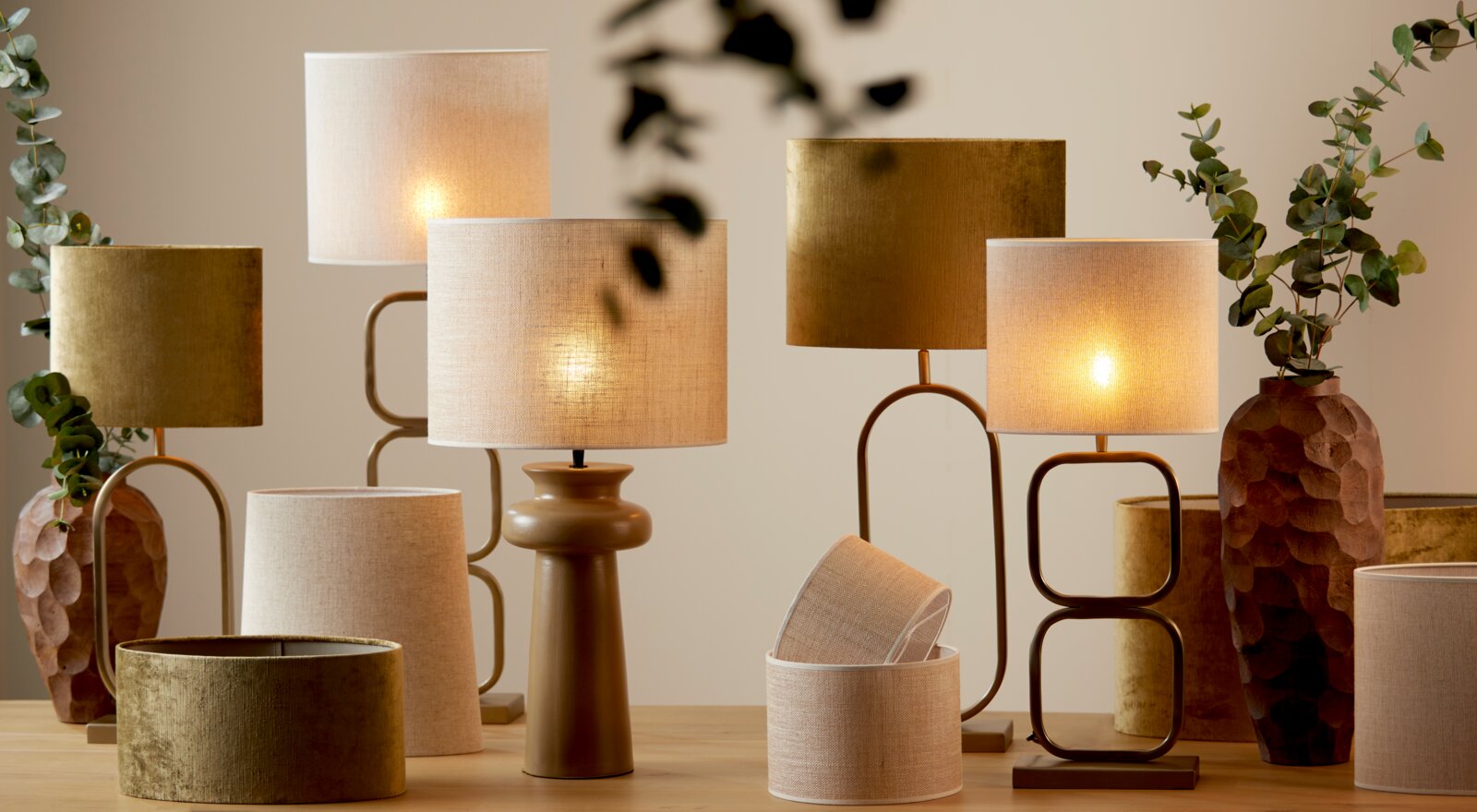 Stimmungsvolle Tischlampen mit warmem Licht und Stoffschirmen in Beige und Oliv auf einem Holztisch, umgeben von dekorativen Vasen und Eukalyptuszweigen; Perspektive: frontale Ansicht auf Augenhöhe.
