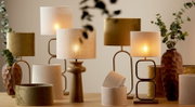 Stimmungsvolle Tischlampen mit warmem Licht und Stoffschirmen in Beige und Oliv auf einem Holztisch, umgeben von dekorativen Vasen und Eukalyptuszweigen; Perspektive: frontale Ansicht auf Augenhöhe.
