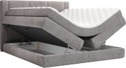 Grijs gestoffeerd boxspringbed met gewatteerd hoofdbord en witte topper, met opklapbare bedbodem die de opbergruimte toont, schuin van voren gezien