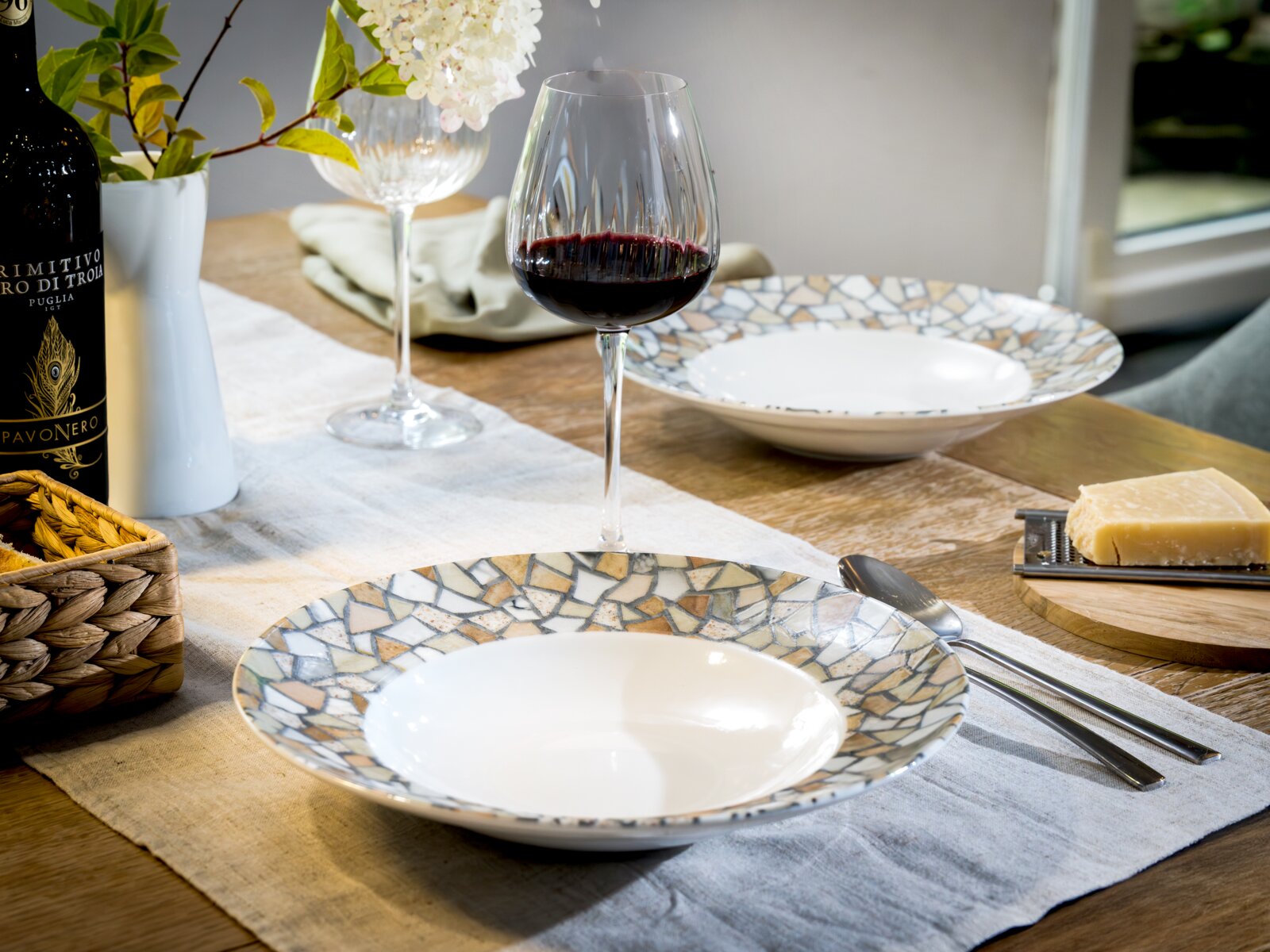 Schräg von vorne fotografierte Tischszene mit zwei dekorativen Keramiktellern im Mosaikdesign, Rotweinglas, Vase mit weißen Blumen, Besteck und Käse auf Holzbrett auf einem Holztisch.