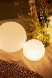 Zwei leuchtende, matte Kugel-Lampen in warmweiß auf einem Tisch, daneben eine grüne Glasvase mit Trockenblumen und Lichterkette; Perspektive: leicht von oben, schräg von vorn.