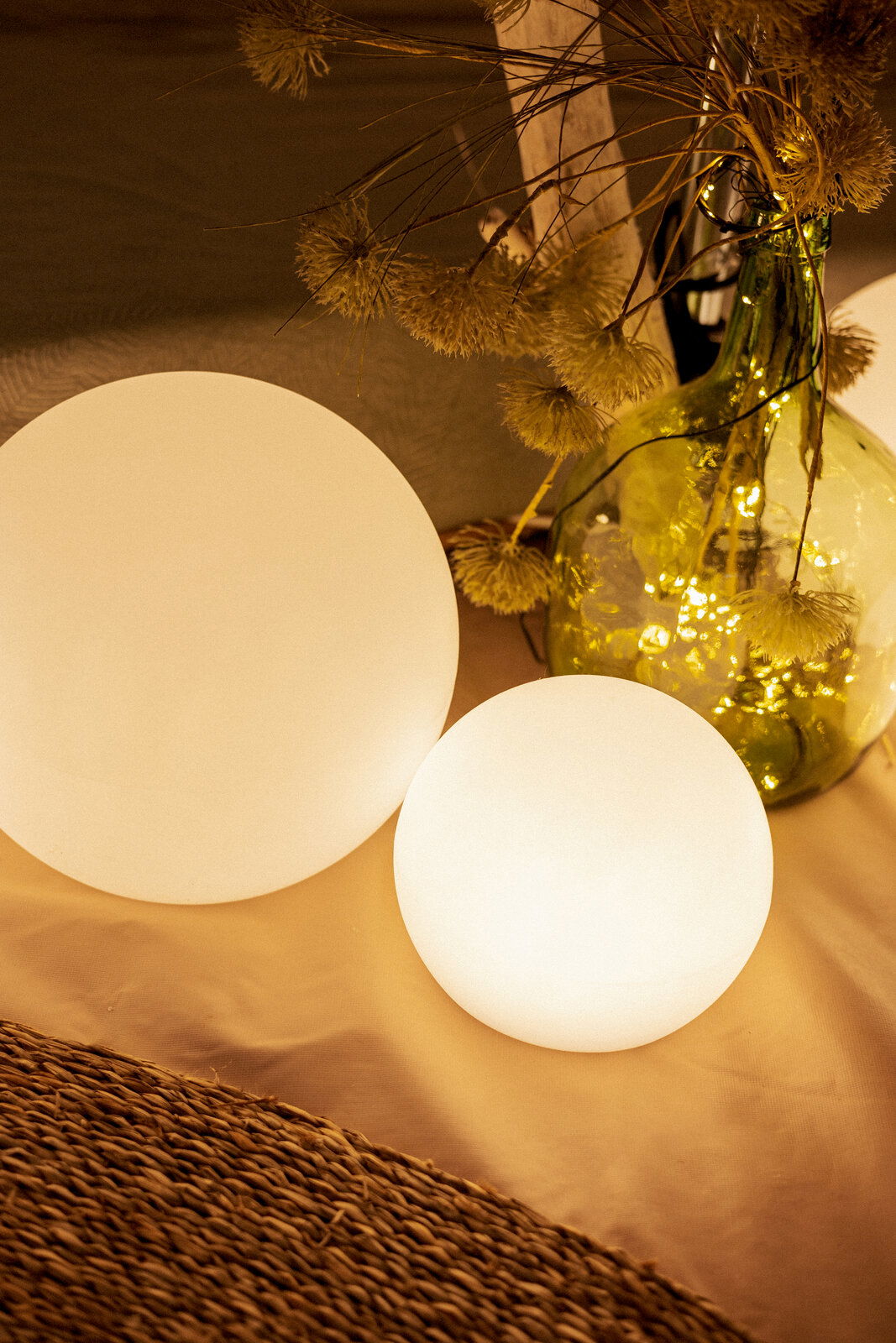 Zwei leuchtende, matte Kugel-Lampen in warmweiß auf einem Tisch, daneben eine grüne Glasvase mit Trockenblumen und Lichterkette; Perspektive: leicht von oben, schräg von vorn.