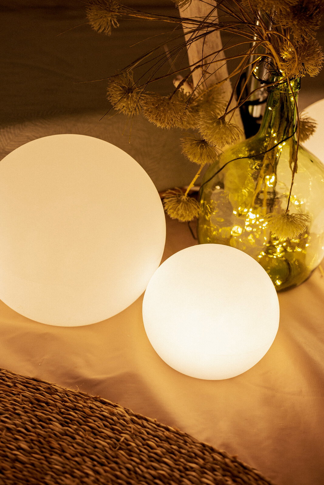 Zwei leuchtende, matte Kugel-Lampen in warmweiß auf einem Tisch, daneben eine grüne Glasvase mit Trockenblumen und Lichterkette; Perspektive: leicht von oben, schräg von vorn.