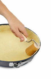 Hand hält einen kleinen Holzteigroller und rollt Teig in einer runden Backform aus, Ansicht von oben.