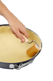 Hand hält einen kleinen Holzteigroller und rollt Teig in einer runden Backform aus, Ansicht von oben.
