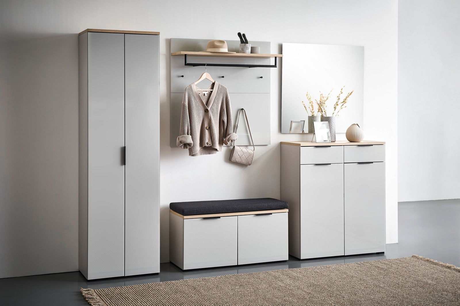 Moderne Garderobenkombination in hellem Raum, bestehend aus einem hohen Schrank, einem Garderobenpaneel mit Haken und Ablage, einer Schuhbank mit Sitzkissen und einem Schuhschrank mit Spiegel. Frontalansicht.