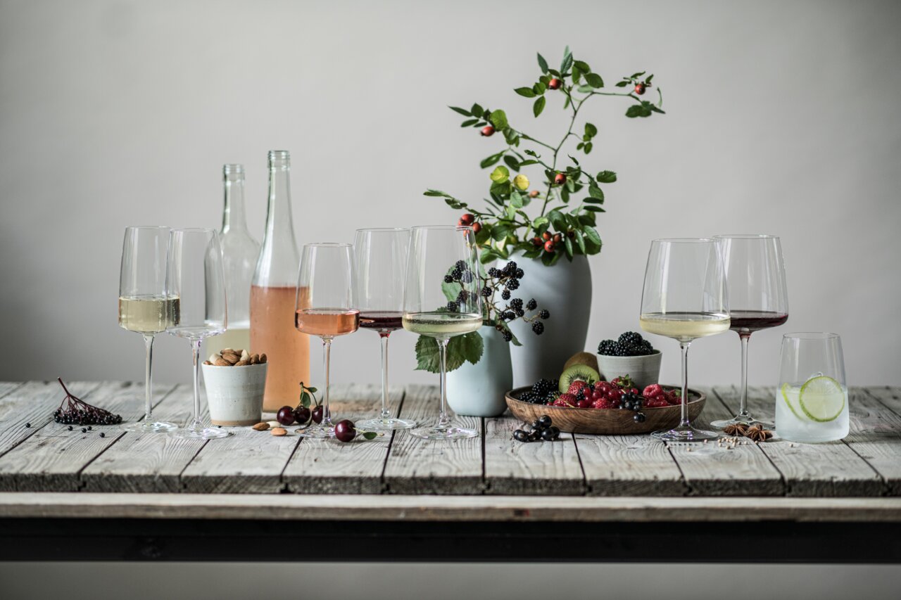 Een stijlvol gedekte tafel met verschillende wijnglazen, gevuld met witte wijn, rosé en rode wijn, omringd door verse bessen, noten en decoratieve planten. Het perspectief toont de glazen vanaf de zijkant.