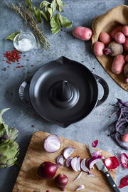 Vogelperspektive auf einen schwarzen STAUB Hähnchenbräter mit zwei Griffen, umgeben von frischen Zutaten wie Kartoffeln, Zwiebeln und Gewürzen auf einem rustikalen Untergrund.