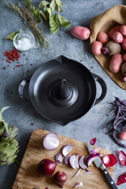 Vogelperspectief op een zwarte STAUB-kippenbraadpan met twee handvatten, omringd door verse ingrediënten zoals aardappelen, uien en kruiden op een rustieke achtergrond.