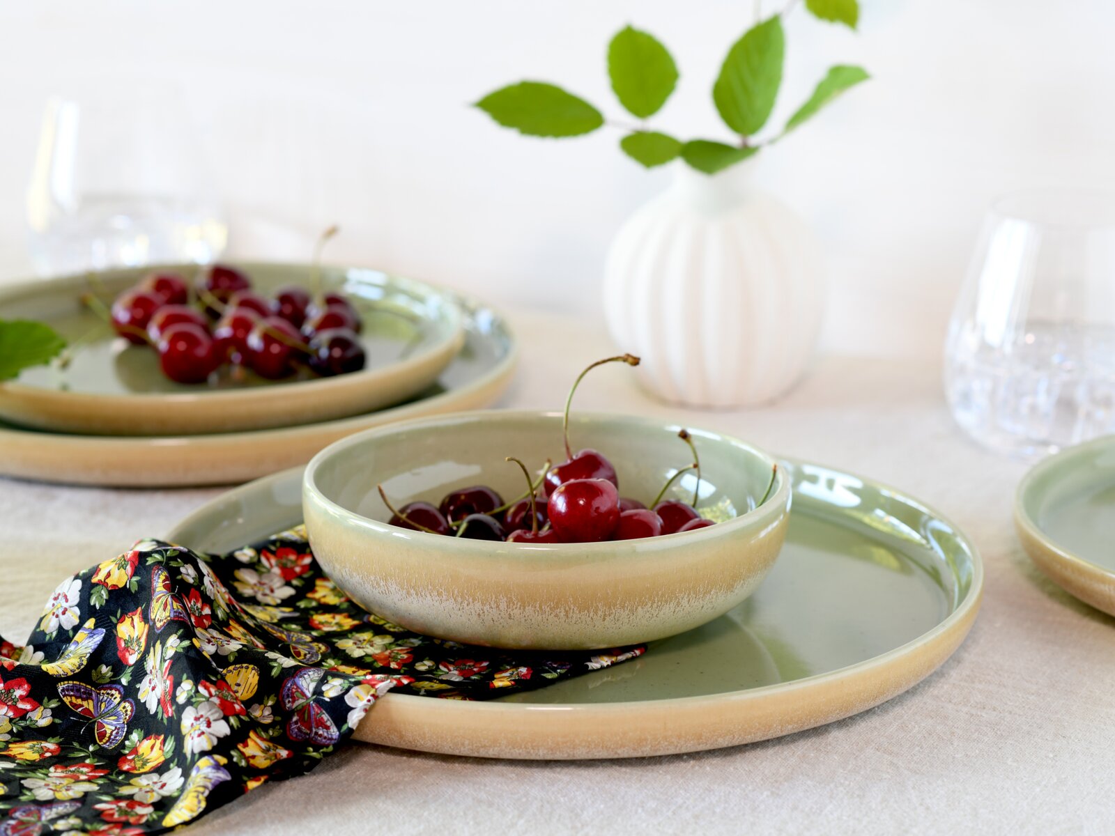 Keramikgeschirr-Set in Salbeigrün und Sandbeige mit großem Teller und Schale, dekoriert mit Kirschen auf einem floralen Tuch; Aufnahme aus leicht schräger Seitenansicht auf Tischhöhe